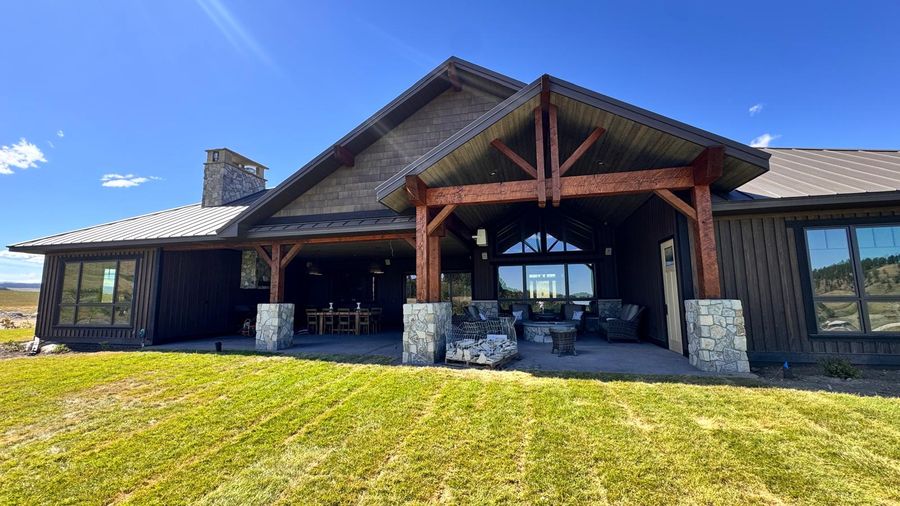 A dark-sided custom home with a timber-framed covered patio and a green lawn